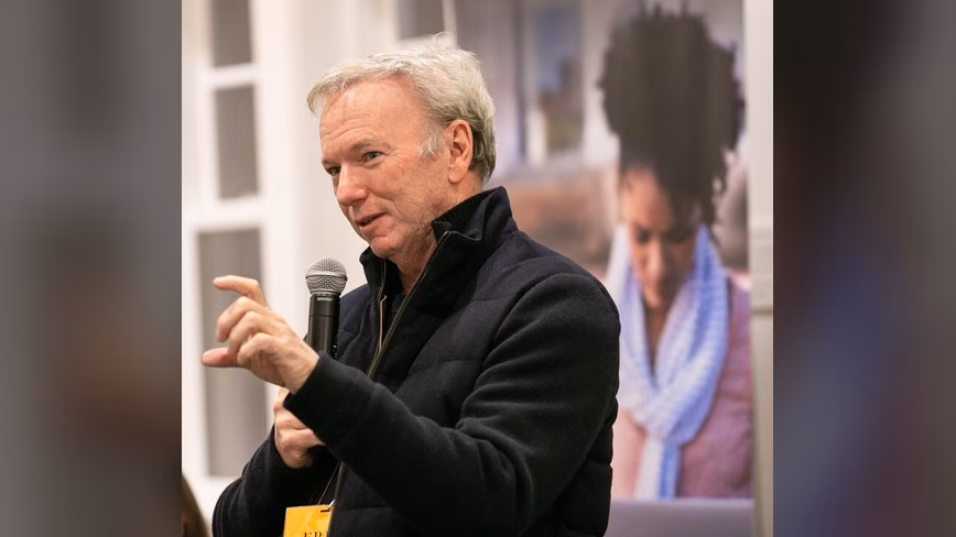 Eric Schmidt, in a dark navy jacket, speaks into a handheld microphone and gestures with his right hand at an indoor event