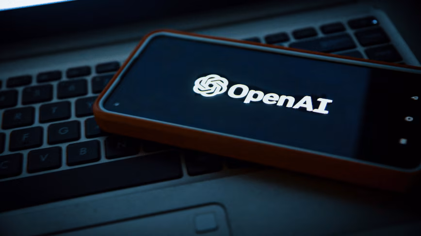 A smartphone in a red case shows the white OpenAI logo on a dark screen, resting on a laptop keyboard in low light