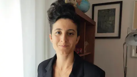 Supplied Sofia has short dark hair that is in a quiff style. She is wearing a black blazer and is standing in a room with a lamp and bookshelf in the background.