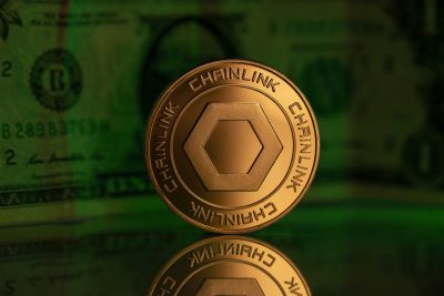 A close-up, eye-level shot of a golden Chainlink cryptocurrency coin standing upright on a highly reflective dark surface. The coin features a prominent hexagonal shape in its center and the word 'CHAINLINK' embossed repeatedly around its edge. In the blurry green background, parts of a U.S. dollar bill are visible, including the number '2' and a serial number 'B 28983769M'. The coin's reflection is clearly visible below it.