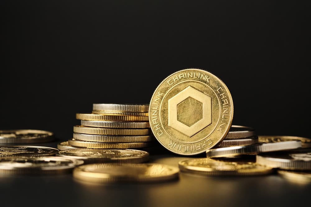 A prominent gold Chainlink cryptocurrency coin, featuring a hexagonal symbol and the word 'CHAINLINK' around its edge, stands upright amidst several stacks and scattered individual gold and silver coins on a dark, reflective surface.