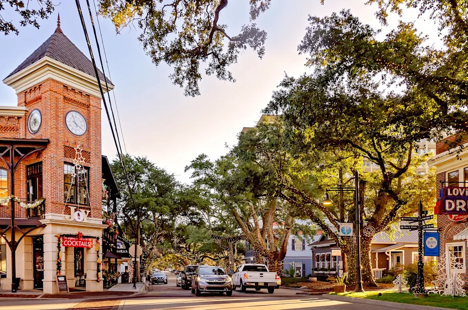 Clock tower in a quaint town square with shops, tree-lined streets, and parked cars under a clear sky