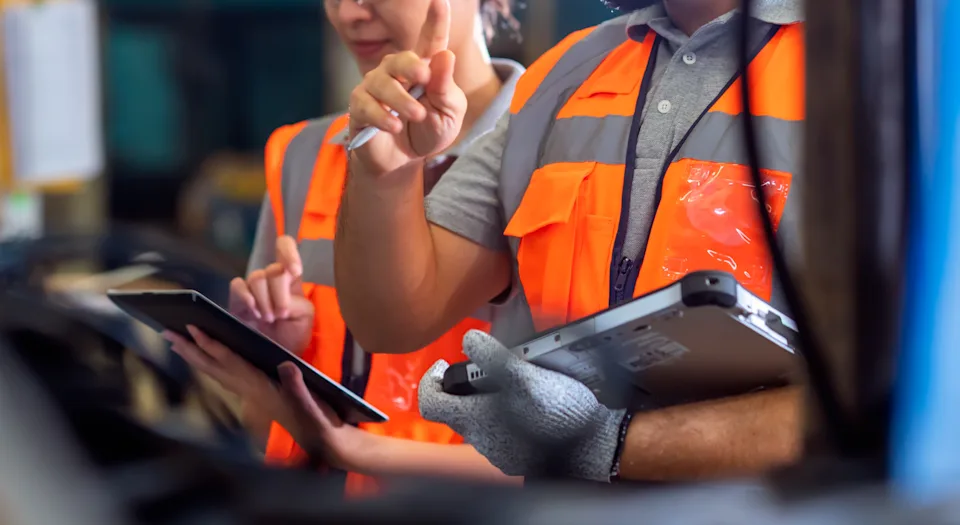 Two construction workers wearing helmets and safety vests discuss work details while holding a tablet and clipboard