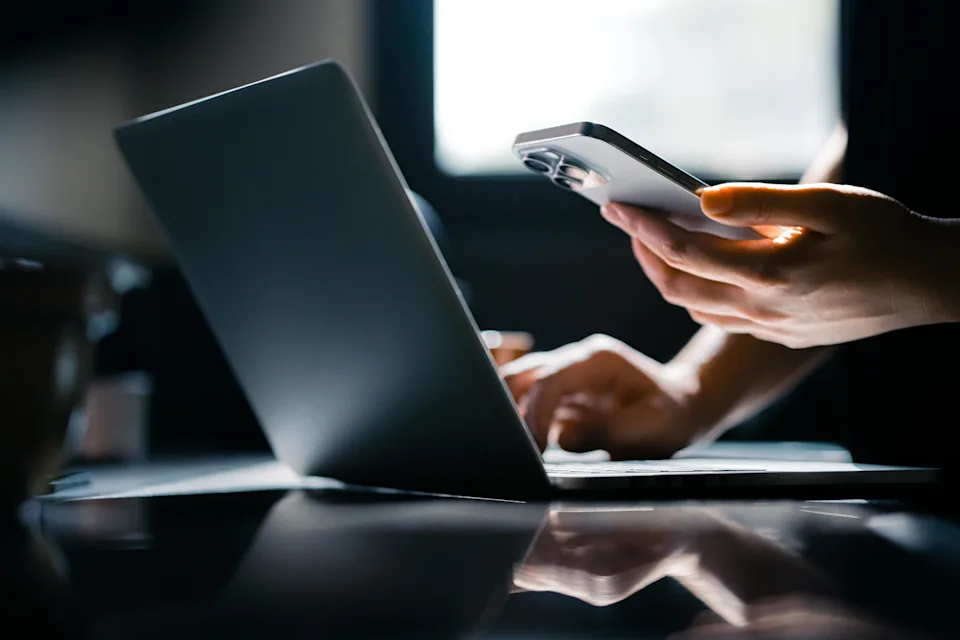 Person using a smartphone and laptop simultaneously, with focus on hands and devices, illustrating multitasking or remote work