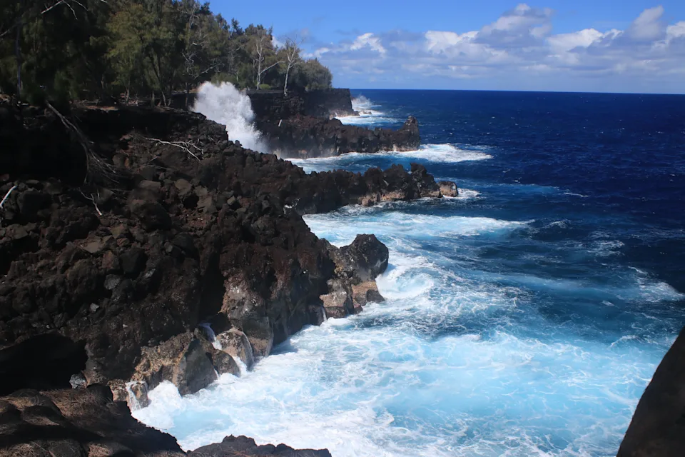 Rocky coastline with waves crashing against the cliffs under a partly cloudy sky. Trees line the edge of the cliffs