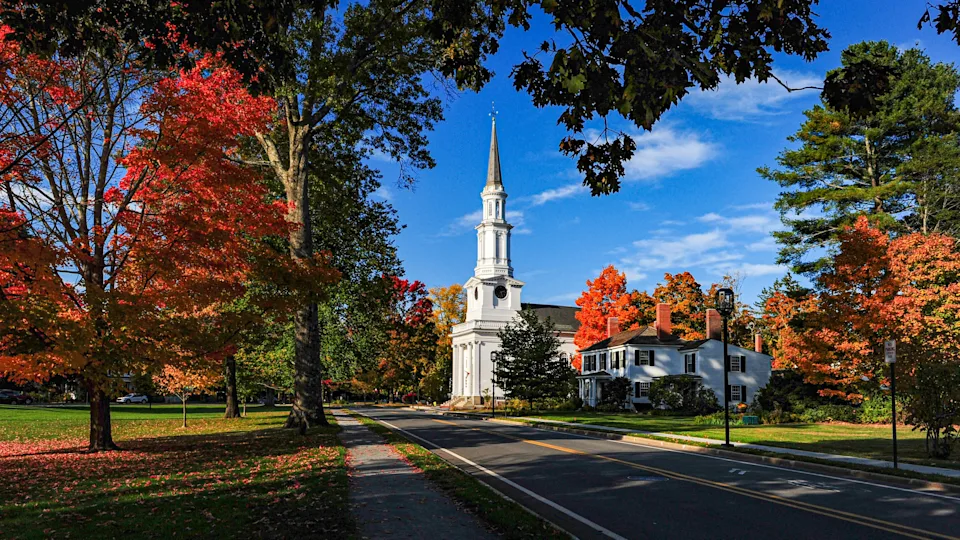 A scenic street with a tall white church surrounded by trees with autumn leaves