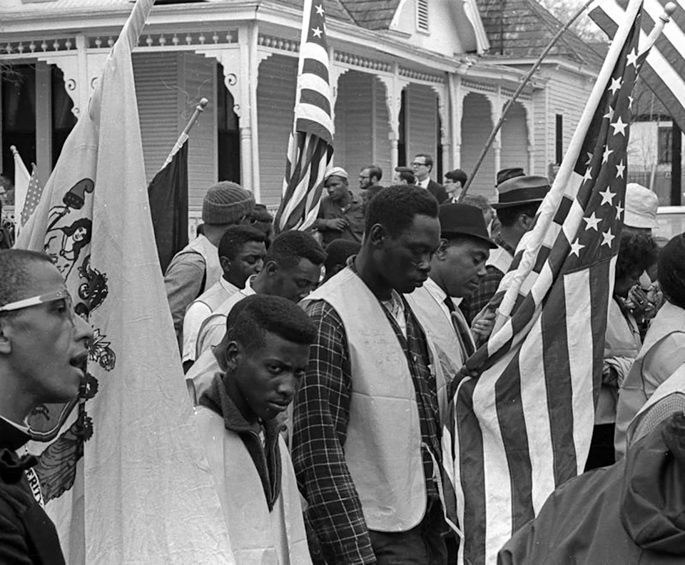 People marching with flags in a civil rights protest, showing determination and unity in front of a traditional house