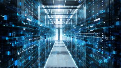 A long, brightly lit hallway inside a data center, flanked by rows of tall server racks on both sides. Bright blue light trails and numerous small blue and white digital squares and dots appear to be moving through the space, emphasizing data flow and connectivity. The perspective looks straight down the aisle towards a bright light at the end.