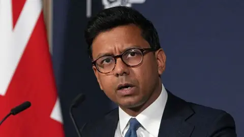 AFP via Getty Images Kanishka Narayan speaking in a suit with a blue tie against a union jack flag backdrop and blue background