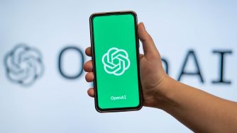 دستی که تلفن هوشمندی با لوگوی OpenAI را در برابر تابلوی محو OpenAI نگه داشته است.