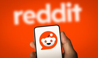 Reddit logo and Reddit mascot displayed on a smartphone held against an orange background.