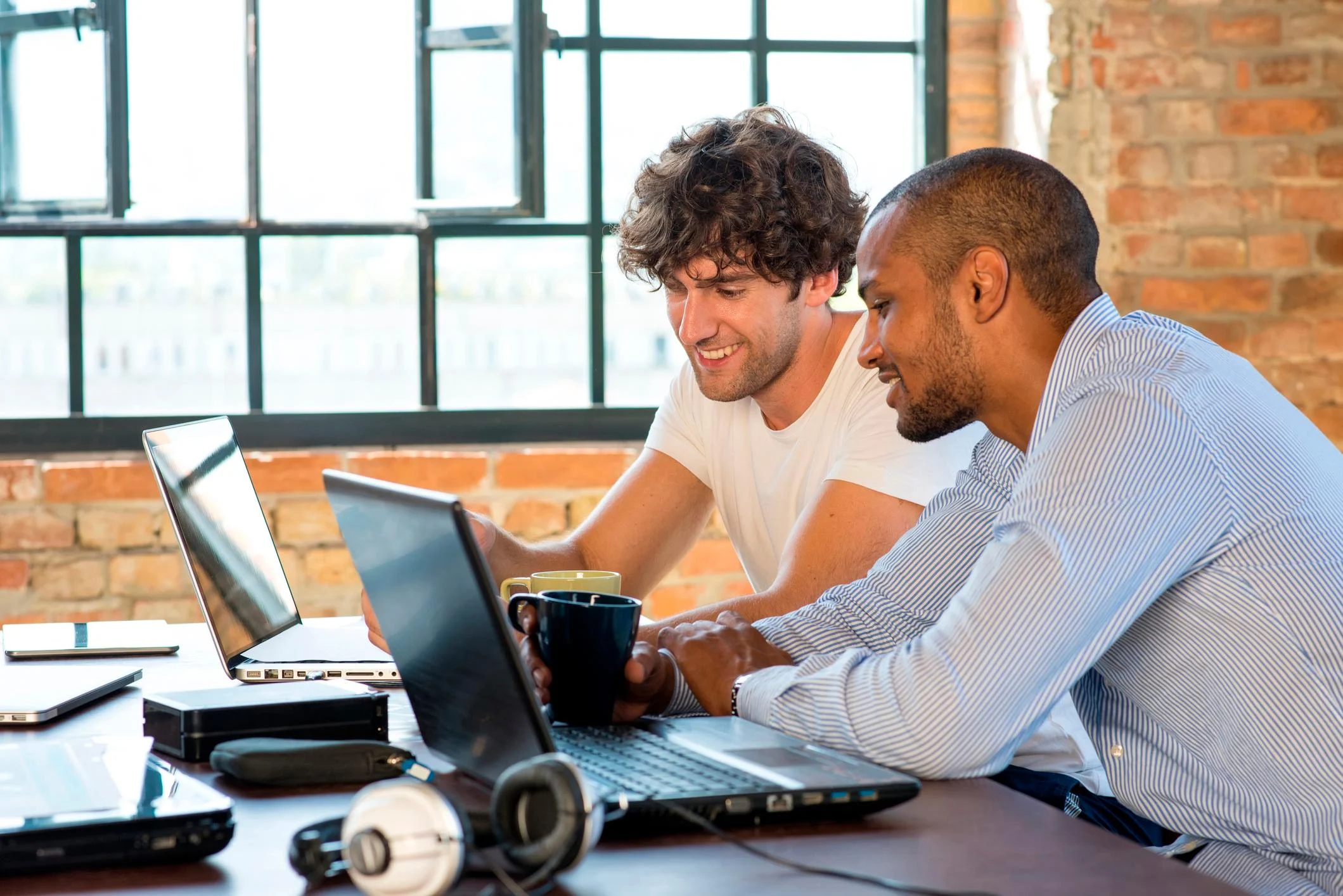 دو همکار در حال همکاری با لپ‌تاپ‌ها در یک دفتر روشن با دیوار آجری