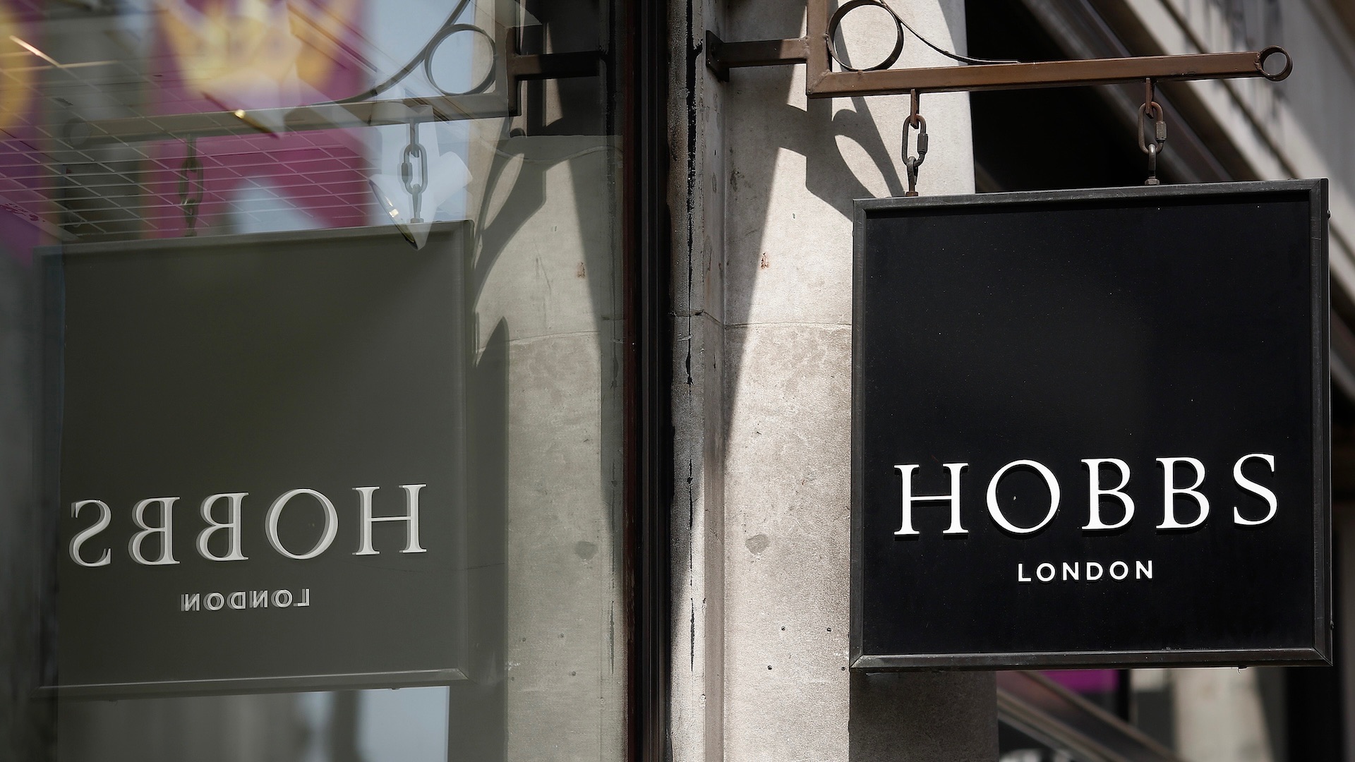 A square, black Hobbs London sign reflected in the window of its store front