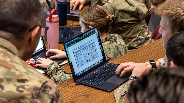 Service members in Army and Navy camouflage uniforms sit at a long wooden table working on laptops. One screen in the foreground shows a research poster.