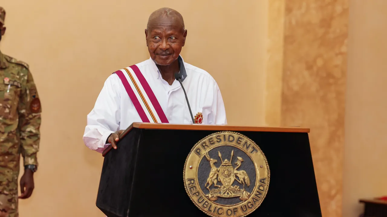 President Yoweri Museveni speaking at a podium bearing the presidential seal during the Independence Day celebration, wearing a white shirt and a maroon, white and gold sash.