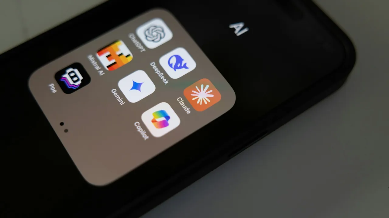 A dark smartphone, seen at an angle, with an open folder labelled AI containing the ChatGPT, Mistral AI, DeepSeek, Poe, Gemini, Claude and Copilot apps