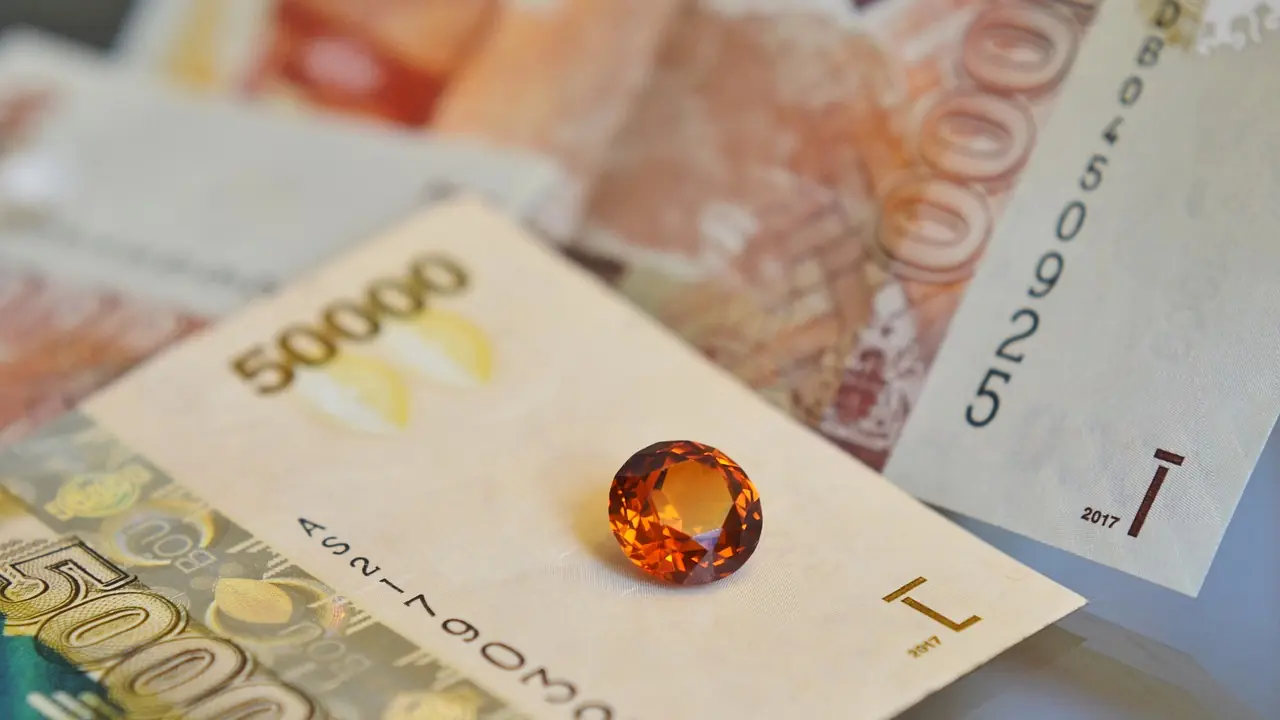 Close-up of a Ugandan 50,000-shilling banknote with an orange round-cut gemstone resting on it, other notes blurred behind