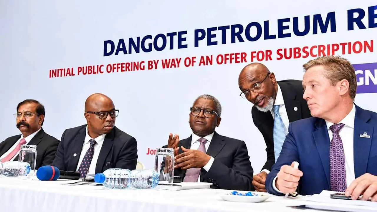 Five men in suits at a table beneath an initial public offering banner, one leaning in to speak to Aliko Dangote, seated centre