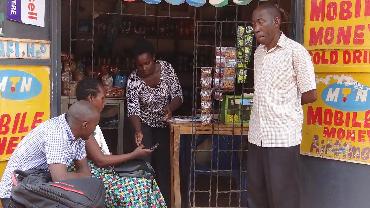 Customers at a roadside kiosk in Uganda covered in yellow MTN Mobile Money signs, one woman showing a phone to the attendant while a man stands nearby.