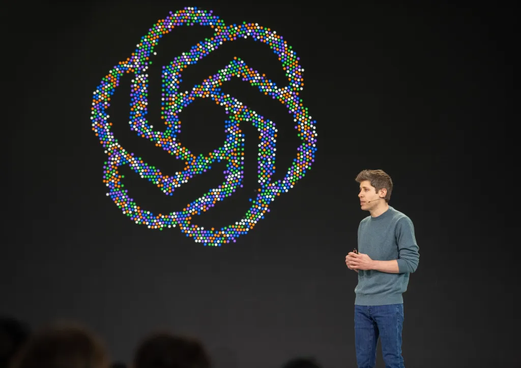Sam Altman speaks on stage at an OpenAI developer conference in front of a large, colorful abstract logo.