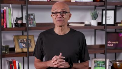 Satya Nadella wears a black t-shirt and glasses, standing with hands clasped in front of a bookshelf decorated with family photos and books, speaking at Microsoft Build 2025.