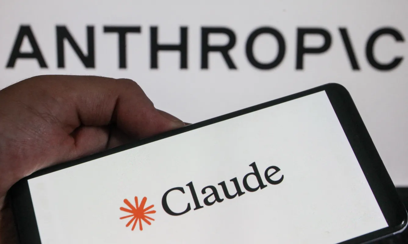 A hand holds a smartphone displaying the logo "Claude" in front of a background with the word "ANTHROPIC."