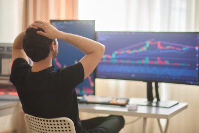 A person, seen from the back, sits in a white chair, hands placed on the back of their head in a gesture of distress or concern. They are looking at two large computer monitors displaying financial candlestick charts with prominent red and green bars, indicating market fluctuations. The charts also show blue and purple trend lines against a dark background. The scene suggests an individual reacting to market performance.