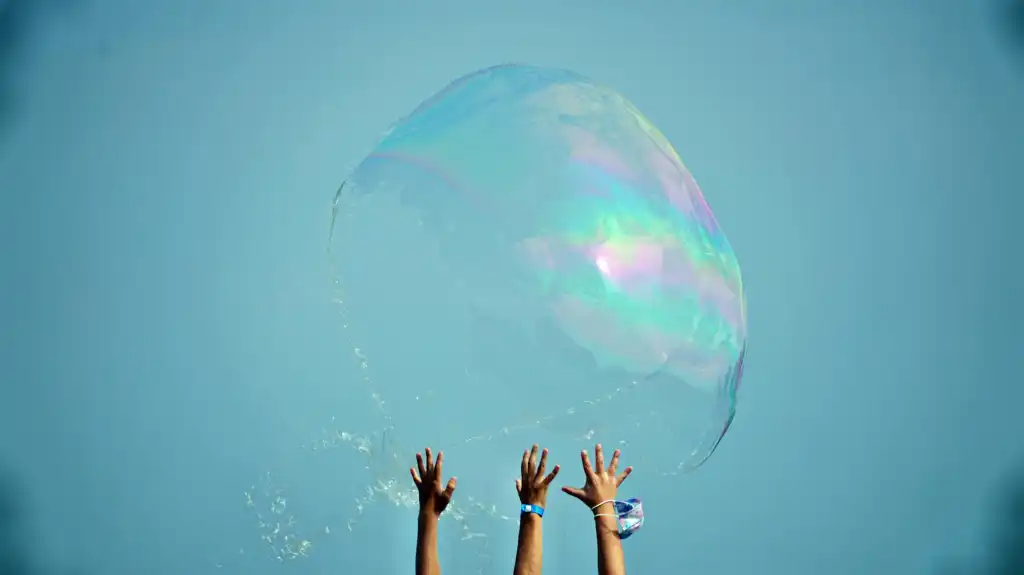 hands of children reach up to giant soap bubble as it pops against blue sky